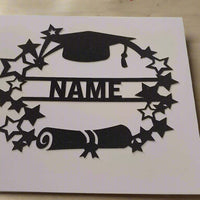 Close-up of a personalised graduation card with black glitter cap, scroll, and star design, photographed from a top-down angle on a wooden surface.