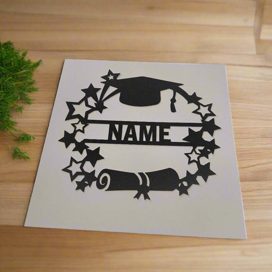 Angled overhead view of a personalised graduation card topper with black glitter mortarboard, stars, and diploma scroll, displayed on a wooden background.