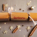 Brown kraft Christmas cracker with white satin ribbon and “Barbara” name in red, styled with cinnamon sticks, star confetti, and warm festive lights.
