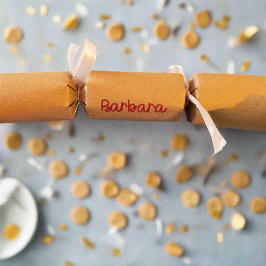 Brown kraft Christmas cracker personalised with “Barbara” in red lettering, tied with white satin ribbon, styled on a table with golden confetti.