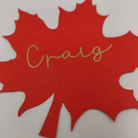 Personalised maple leaf name card cut from red cardstock with gold handwritten script reading Craig, perfect for autumn or Thanksgiving table settings.