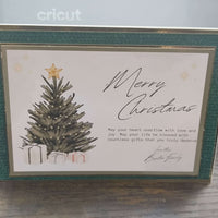 Handmade luxury Christmas card standing upright on a wooden surface. The card features a traditional green textured border with gold foil accents, a central illustration of a Christmas tree topped with a gold star, surrounded by presents. The text reads “Merry Christmas” in elegant script, followed by a heartfelt message and signed “Love the Baxter Family.”