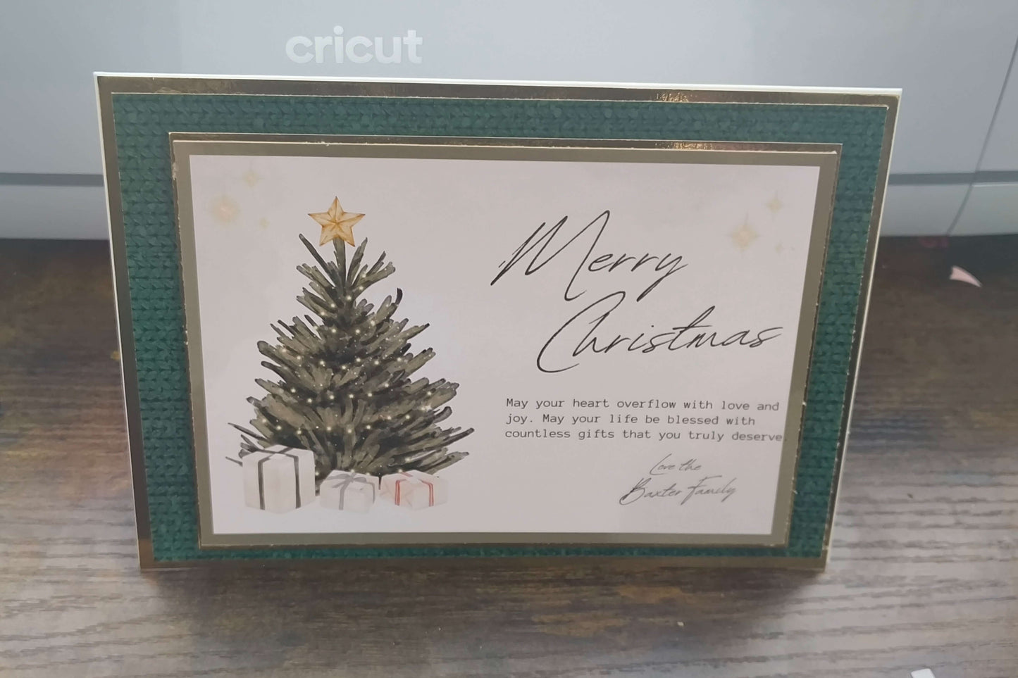 Handmade luxury Christmas card standing upright on a wooden surface. The card features a traditional green textured border with gold foil accents, a central illustration of a Christmas tree topped with a gold star, surrounded by presents. The text reads “Merry Christmas” in elegant script, followed by a heartfelt message and signed “Love the Baxter Family.”