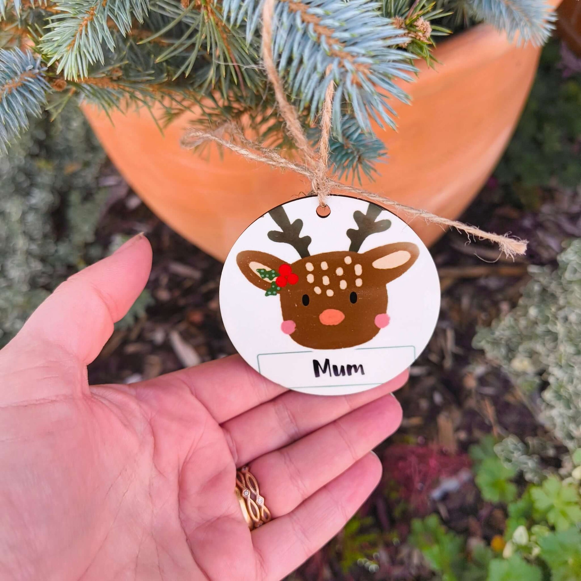 Hand holding personalised reindeer Christmas bauble with ‘Mum’ text, rustic jute twine, and fir tree background