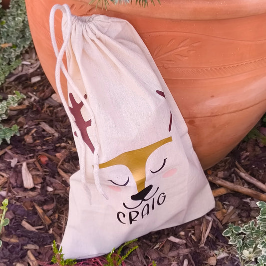 Personalised drawstring gift bag with reindeer face design, name Craig, placed against terracotta plant pot outdoors