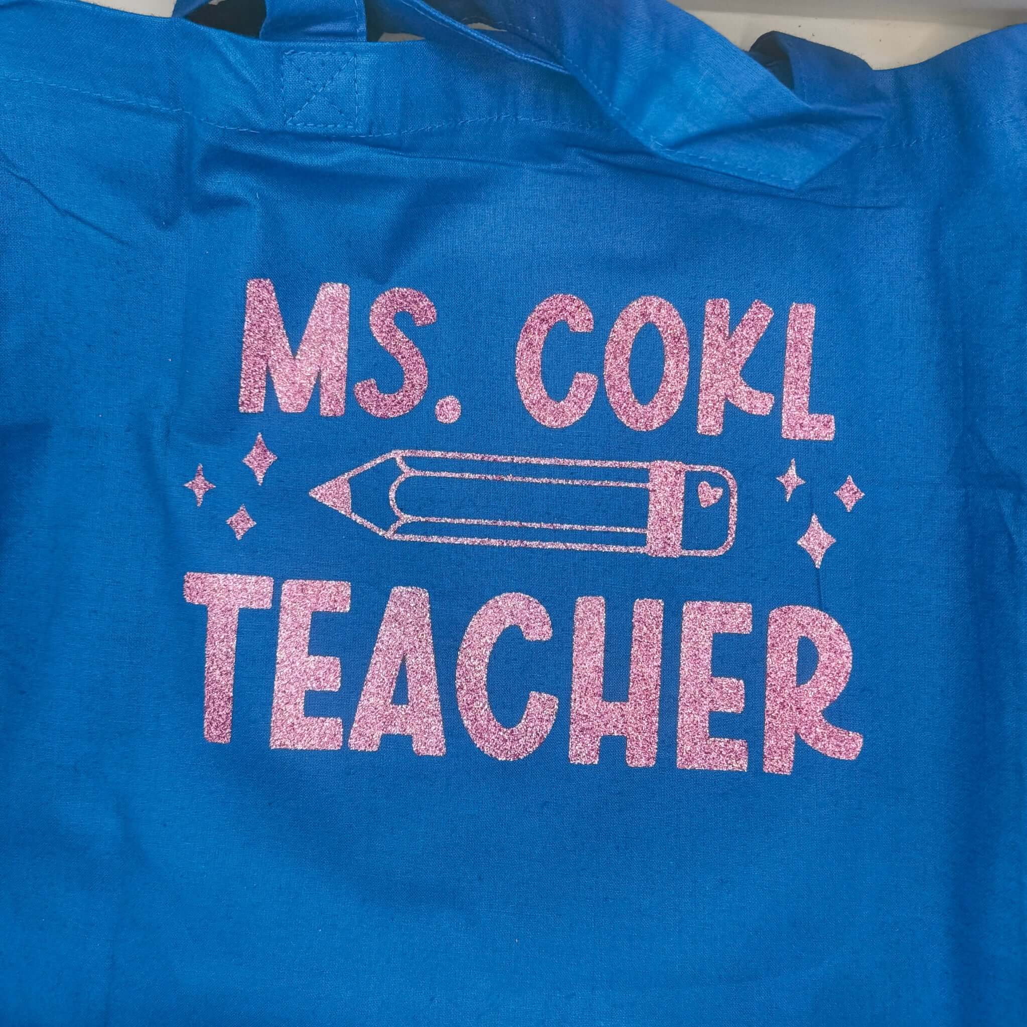 Blue tote bag with pink glitter vinyl text "Ms. Cohl Teacher" featuring a pencil illustration, placed on a wooden surface in a bright home setting.