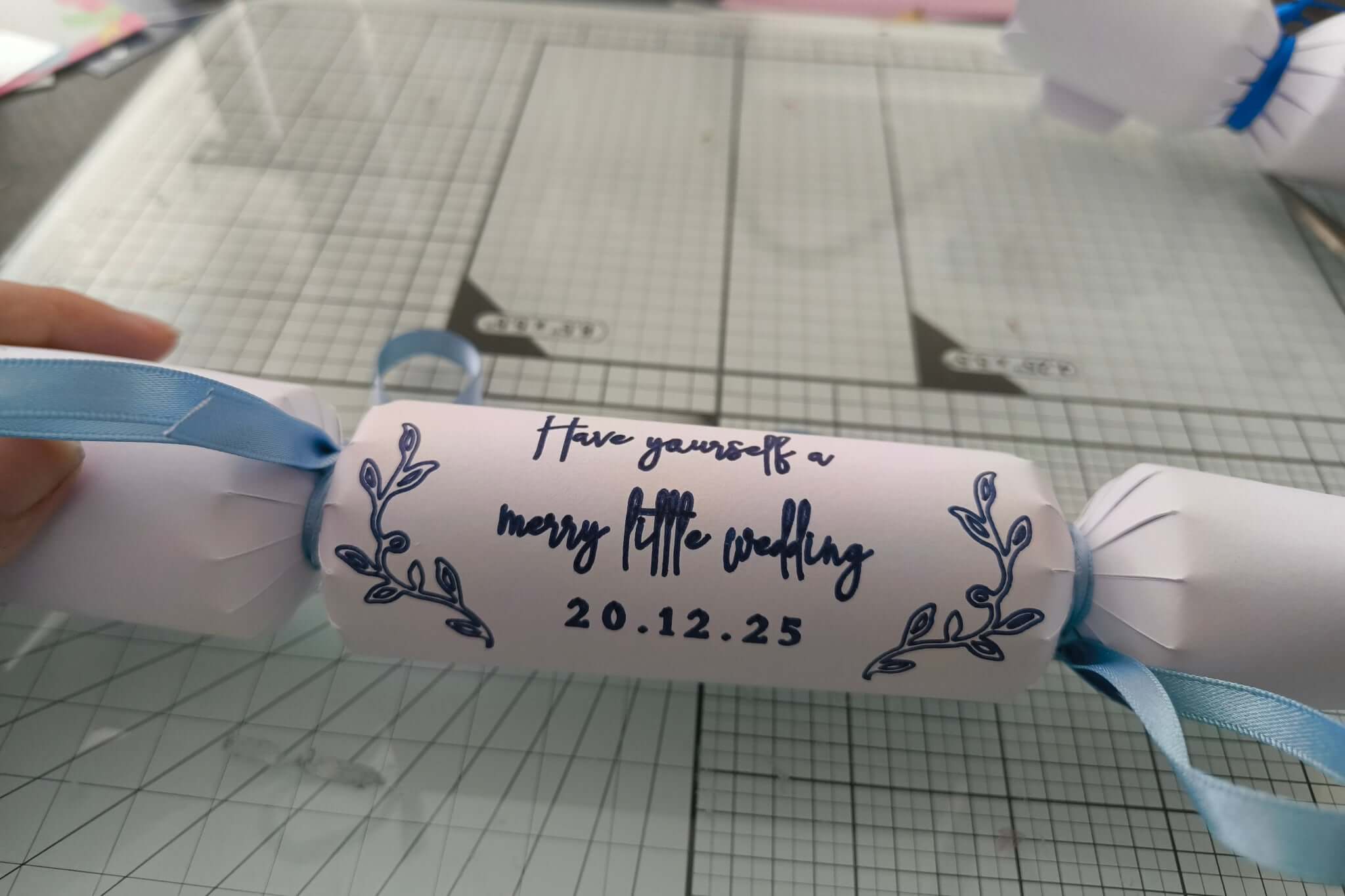 Close-up of white cracker with navy foil wedding message and date, tied with light blue satin ribbon, placed on craft grid surface.
