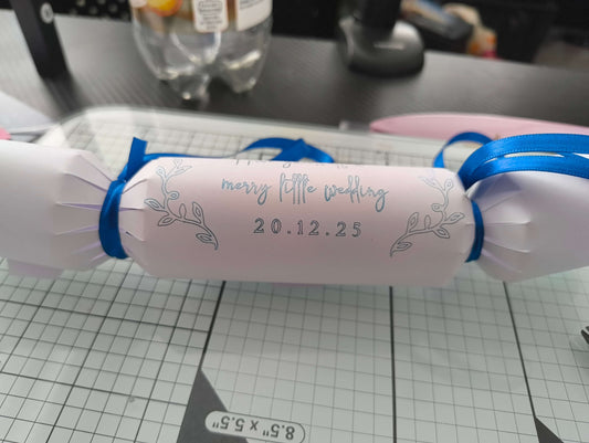 White wedding cracker with silver foil lettering and date, finished with bright blue-ribbon bows, placed on a cutting mat.