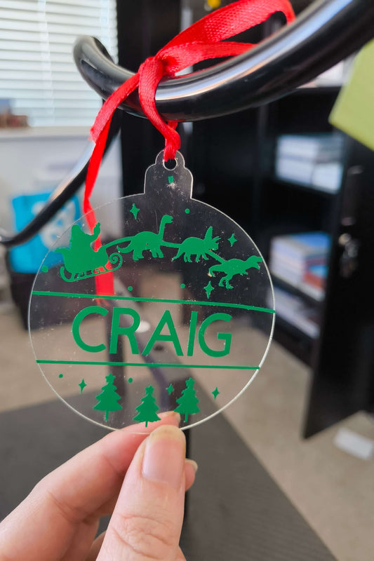 Clear acrylic dinosaur Christmas bauble with green vinyl, personalised with the name Craig, Santa in sleigh pulled by dinosaurs.