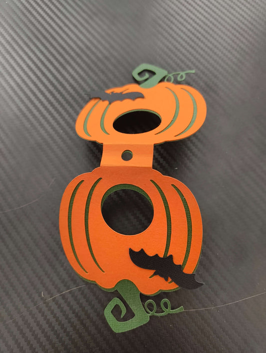 Front close-up of a handmade pumpkin-shaped lollipop holder in bright orange with black bat detail and green stem, photographed from above on a black carbon-textured background.