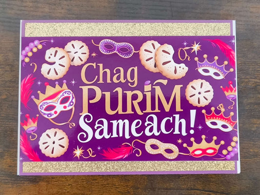 Top-down view of handmade Chag Purim Sameach card with gold glitter trim, purple background, hamantaschen, masks, crowns, and feathers in bold festive colours