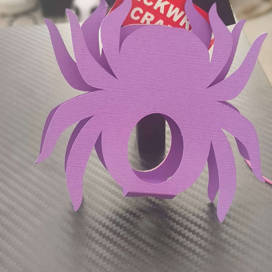 Back view of a handmade purple spider-shaped lollipop holder made from textured cardstock, displayed on a dark carbon-look surface, ideal for Halloween crafts or party favours.