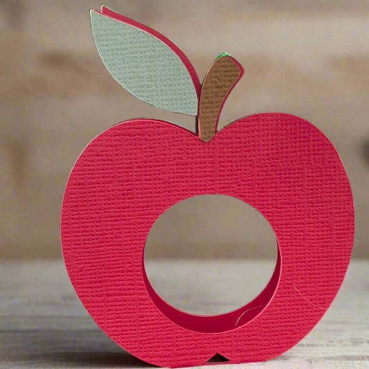 Close-up of a handmade red apple-shaped lollipop holder made from textured cardstock with a brown stem and green leaf, shown upright against a dark background.