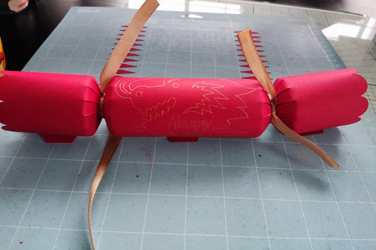 Handmade red Chinese New Year cracker with gold foil dragon design, tied with gold ribbon, shown flat on a crafting mat during assembly.