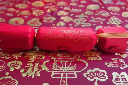Red cracker with gold foil “Happy Chinese New Year” lettering, tied with gold ribbon, displayed on a red and gold patterned festive backdrop with traditional Chinese motifs.