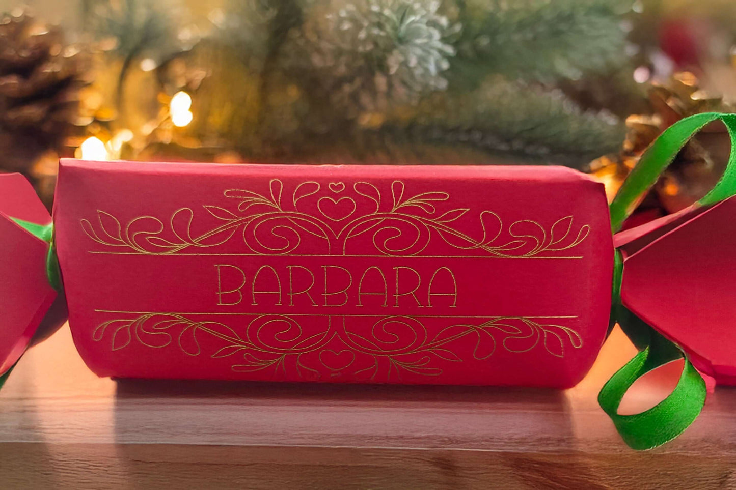 Red Christmas cracker with gold foil personalised name “Barbara” and ornate swirl design, tied with green satin ribbon, styled on wood with festive pinecones and lights.