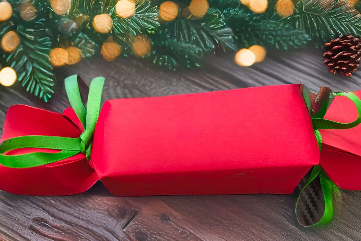 Plain red cracker tied with green ribbon, displayed on rustic wooden table with pinecones and festive garland with warm golden lights.