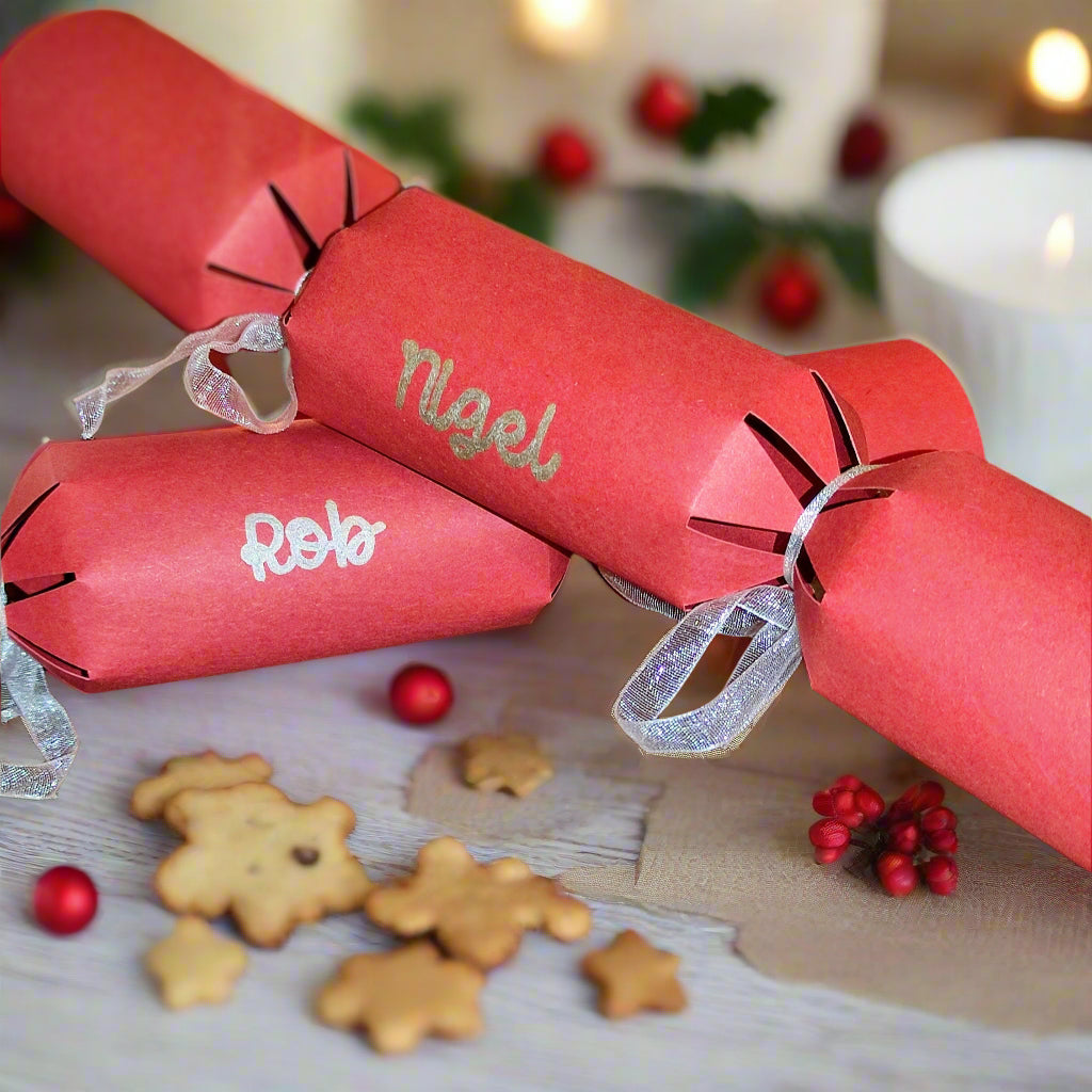 Red personalised Christmas crackers with names “Nigel” and “Rob” in silver lettering, decorated with silver ribbon, styled with gingerbread biscuits and festive berries.
