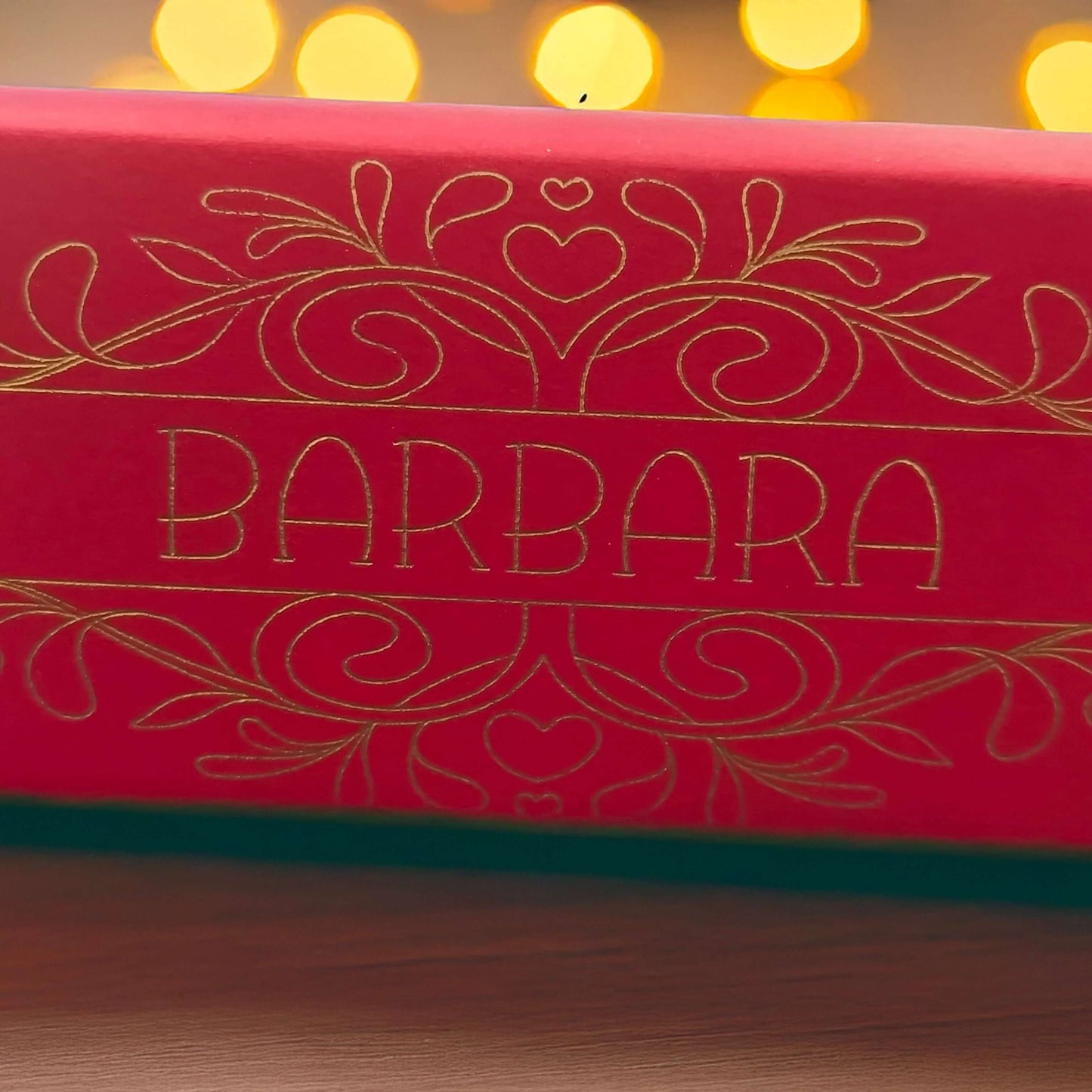 Close-up of red cracker with intricate gold foil design and name “Barbara,” captured with warm bokeh lights in the background for a festive glow.