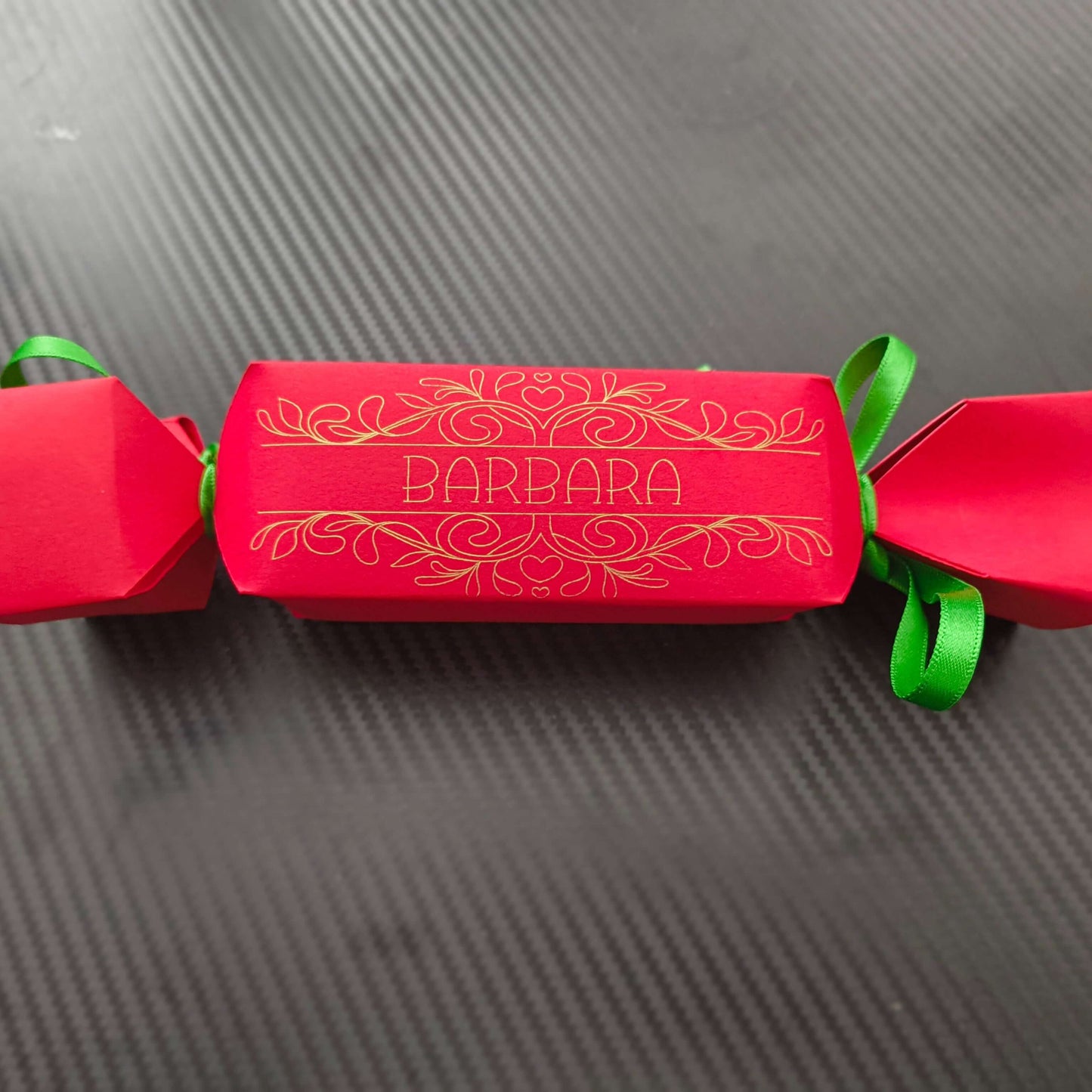 Red cracker with gold foil design and name “Barbara,” tied with green ribbon, photographed flat on textured black background for modern contrast.