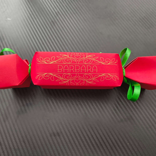 Red cracker with gold foil design and name “Barbara,” tied with green ribbon, photographed flat on textured black background for modern contrast.