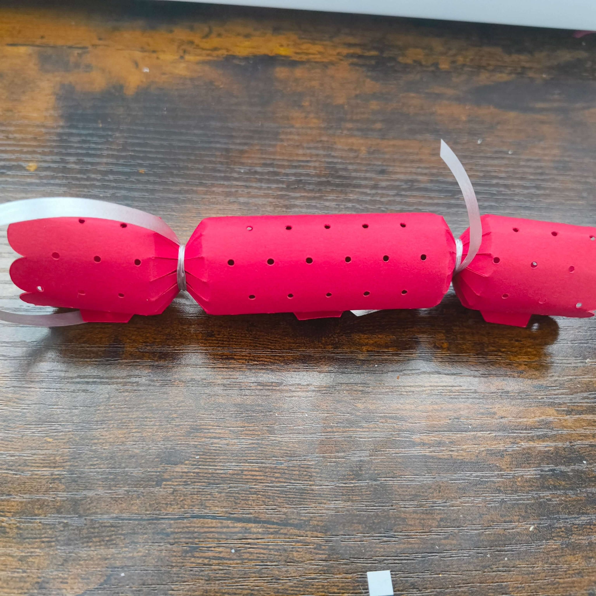 Plain red Christmas cracker with dotted cut-out design and scalloped edges, tied with silver ribbon, placed on wooden tabletop.