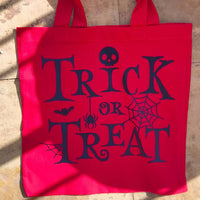 Red trick or treat tote bag laid flat outdoors, showing black Halloween lettering with spider, skull and cobweb design.