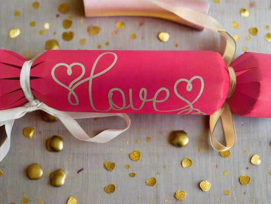 Close-up of a red cracker with “Love” in gold pen, tied with ribbon and surrounded by golden heart confetti, perfect for weddings or Valentine’s.