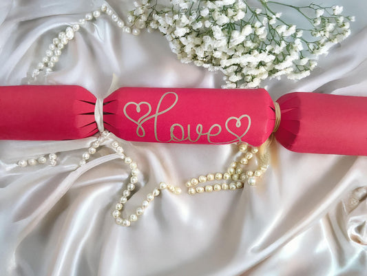 A red cracker with “Love” hand-drawn in gold pen, tied with white ribbon, styled on satin with pearls and white flowers for a romantic look.