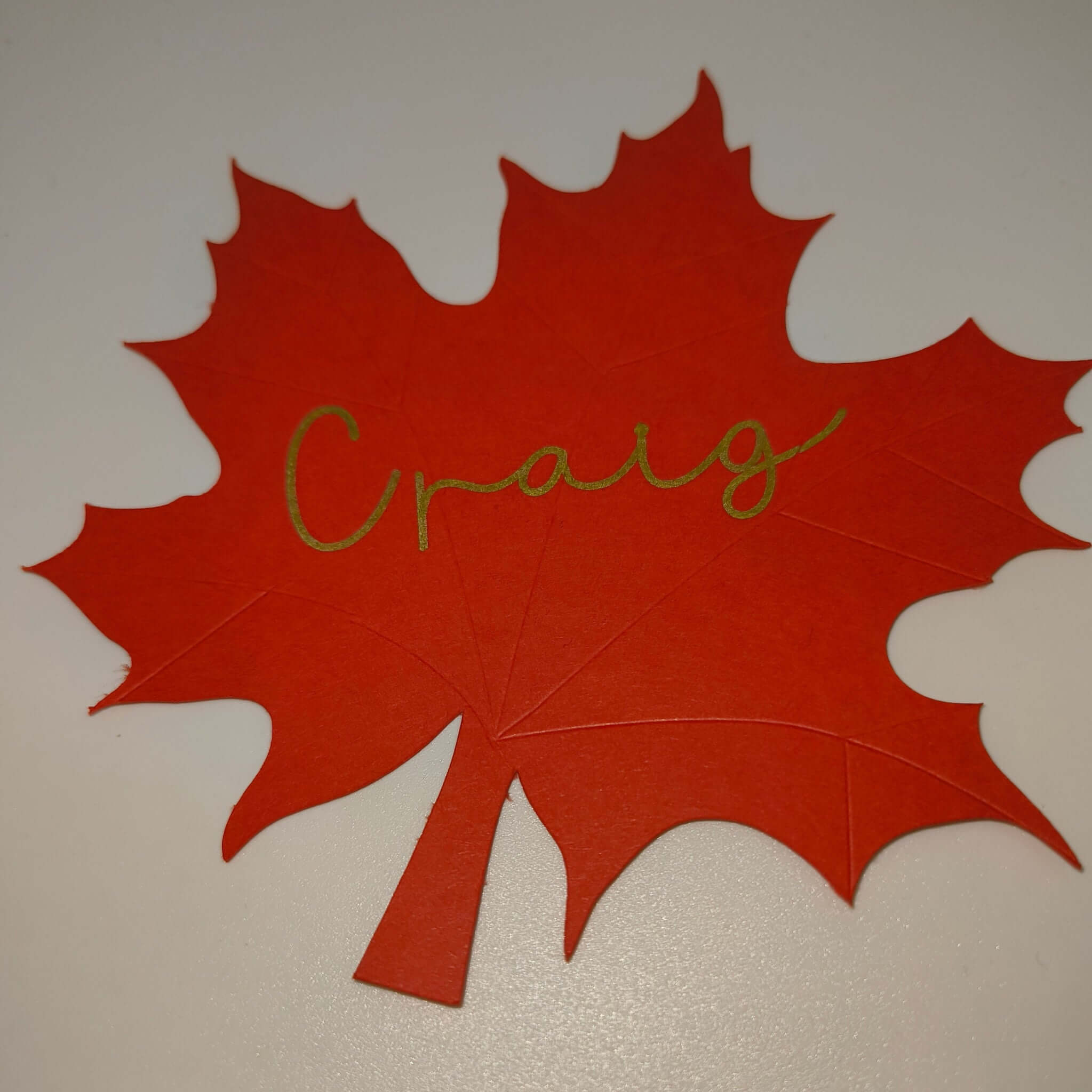Red maple leaf place card with the name Craig written in gold script, handmade from cardstock, shown on a white background.