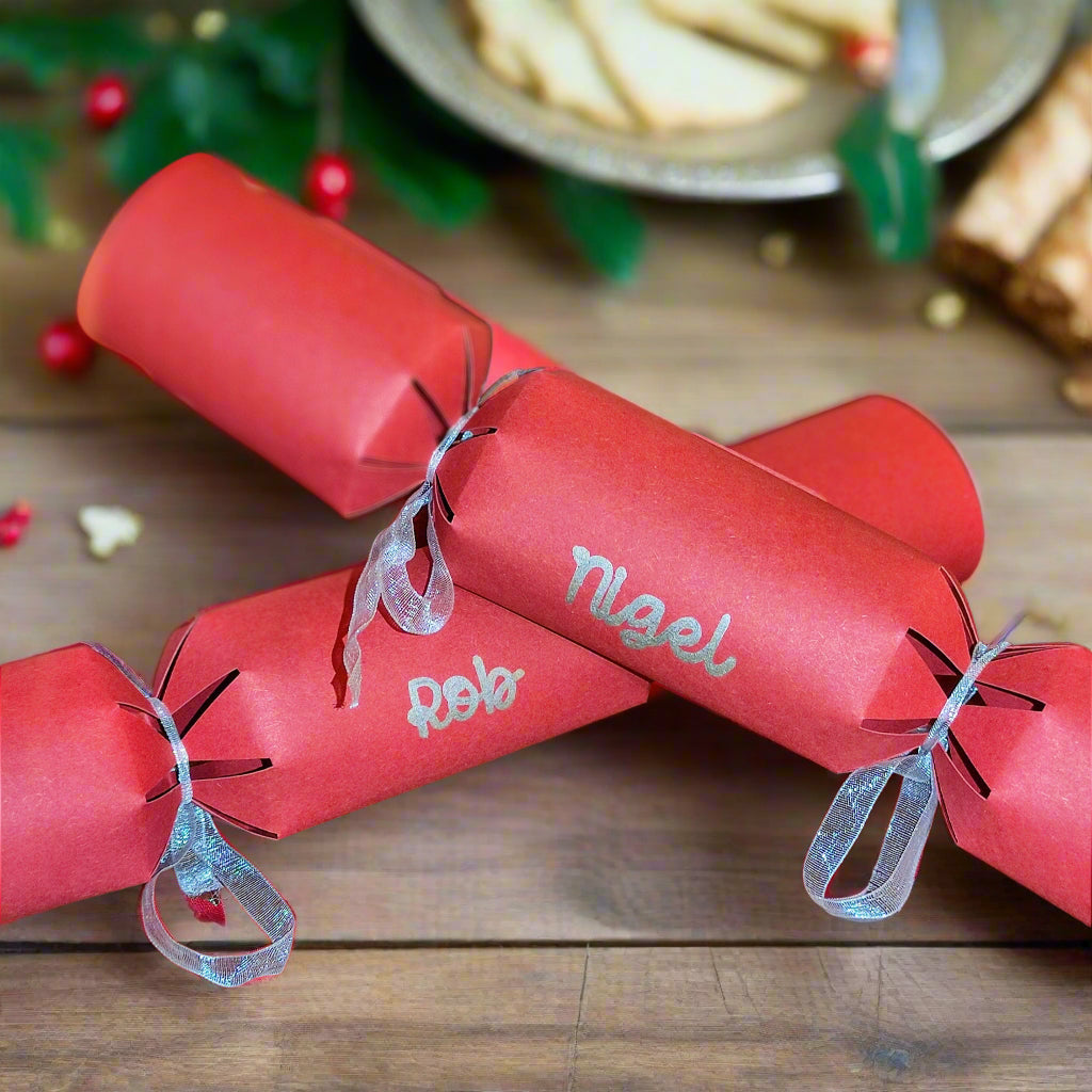 Set of red Christmas crackers with names “Nigel” and “Rob” in glitter lettering, tied with silver ribbon, styled with festive greenery on a wooden table.
