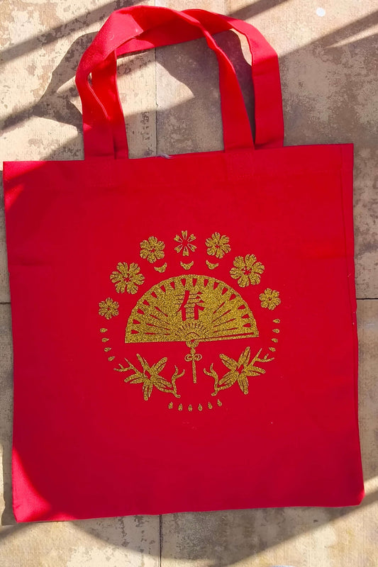 Back view of red tote bag with gold glitter fan and floral vinyl design, laid flat on stone background.