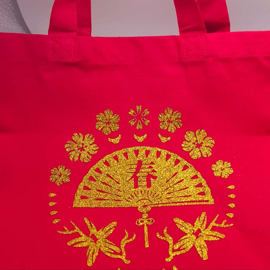 Red fabric tote bag with sturdy handles, featuring a glitter gold fan and floral design at the centre.