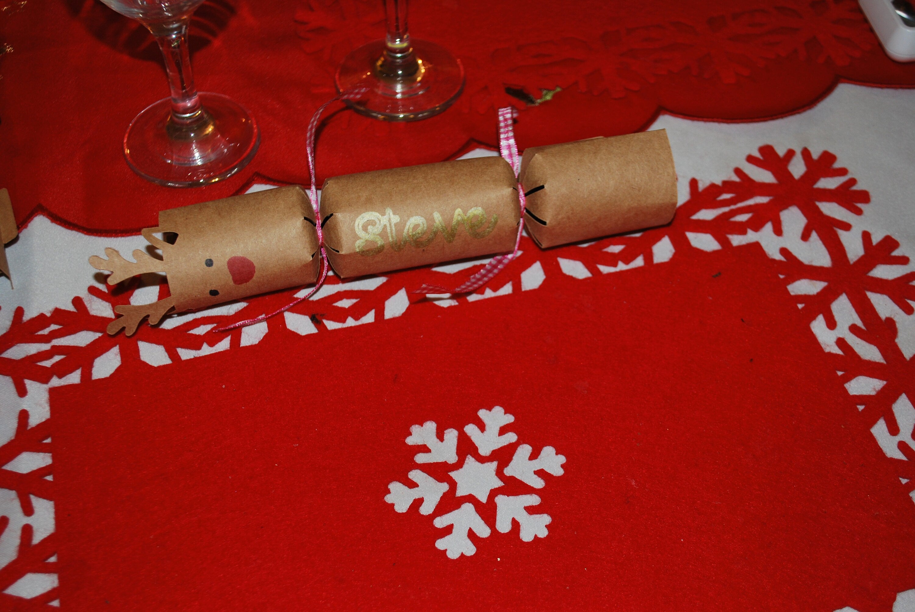 Single kraft reindeer cracker with antlers, red nose, and “Steve” in gold lettering, tied with pink ribbon and placed on a red snowflake placemat.