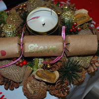 Handmade reindeer cracker in brown kraft card with red nose, black eyes, and “Steve” in gold lettering, tied with pink gingham ribbon, styled on a festive wreath with candle.