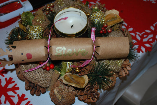 Handmade reindeer cracker in brown kraft card with red nose, black eyes, and “Steve” in gold lettering, tied with pink gingham ribbon, styled on a festive wreath with candle.