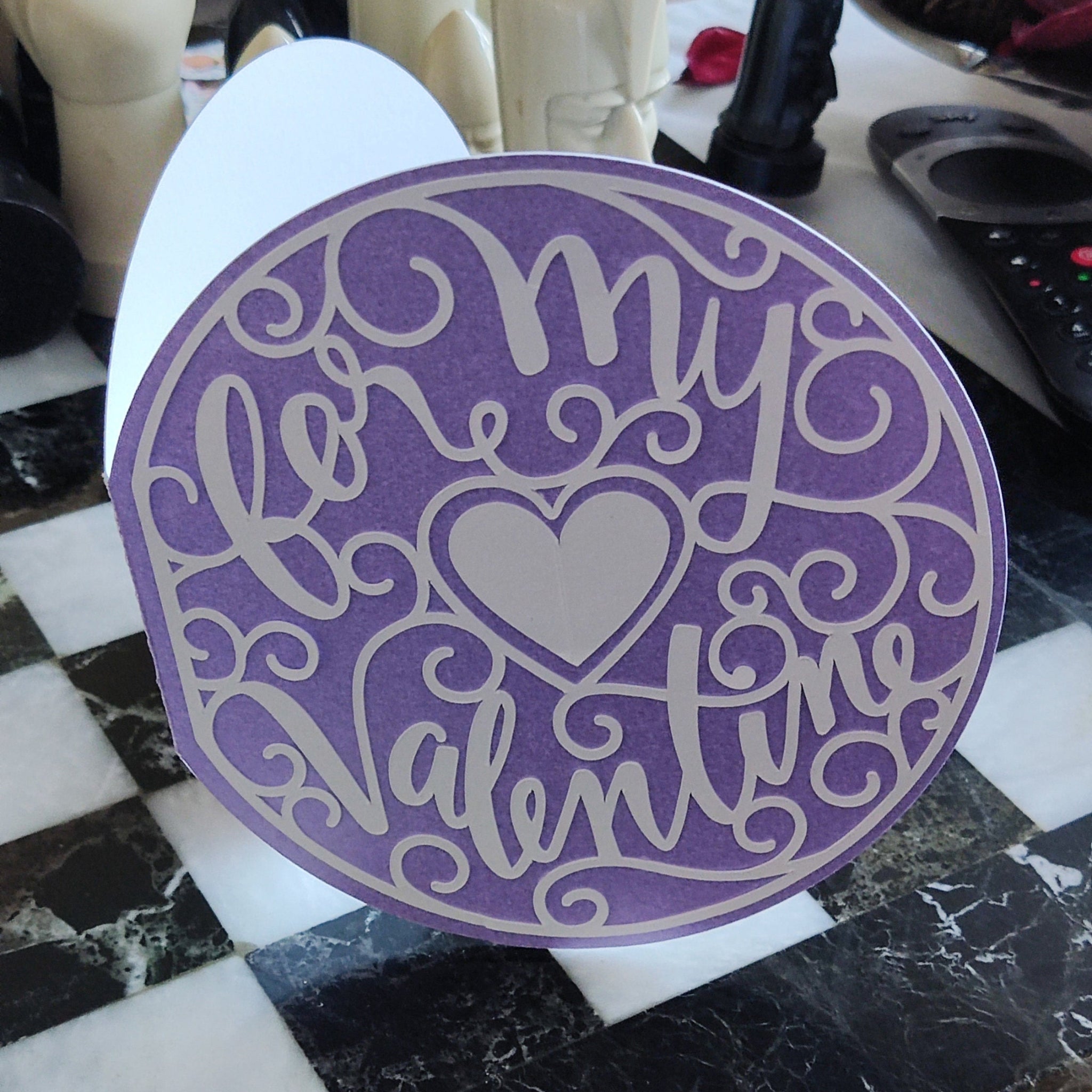 Standing round Valentine’s card in lilac with cream cut-out lettering and heart motif, displayed on a checkered tabletop with objects in the background for scale.
