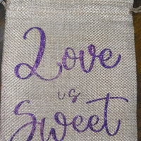 Close-up of a hessian favour bag with shimmering purple glitter lettering “Love is Sweet.” Woven, textured finish with drawstring closure, perfect for weddings or gifts.