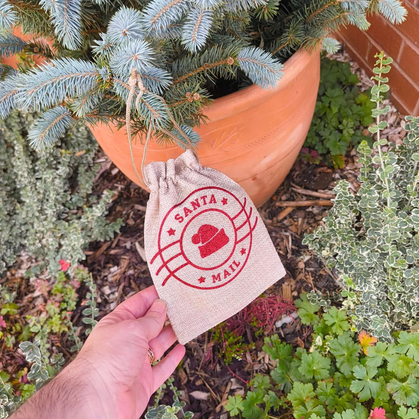 Close-up of Santa Mail hessian drawstring bag with red festive design against pine tree background.