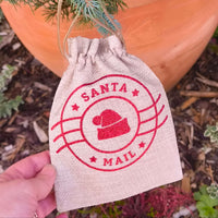 Hand holding a small hessian Santa Mail gift bag with red design, outdoors near a Christmas tree