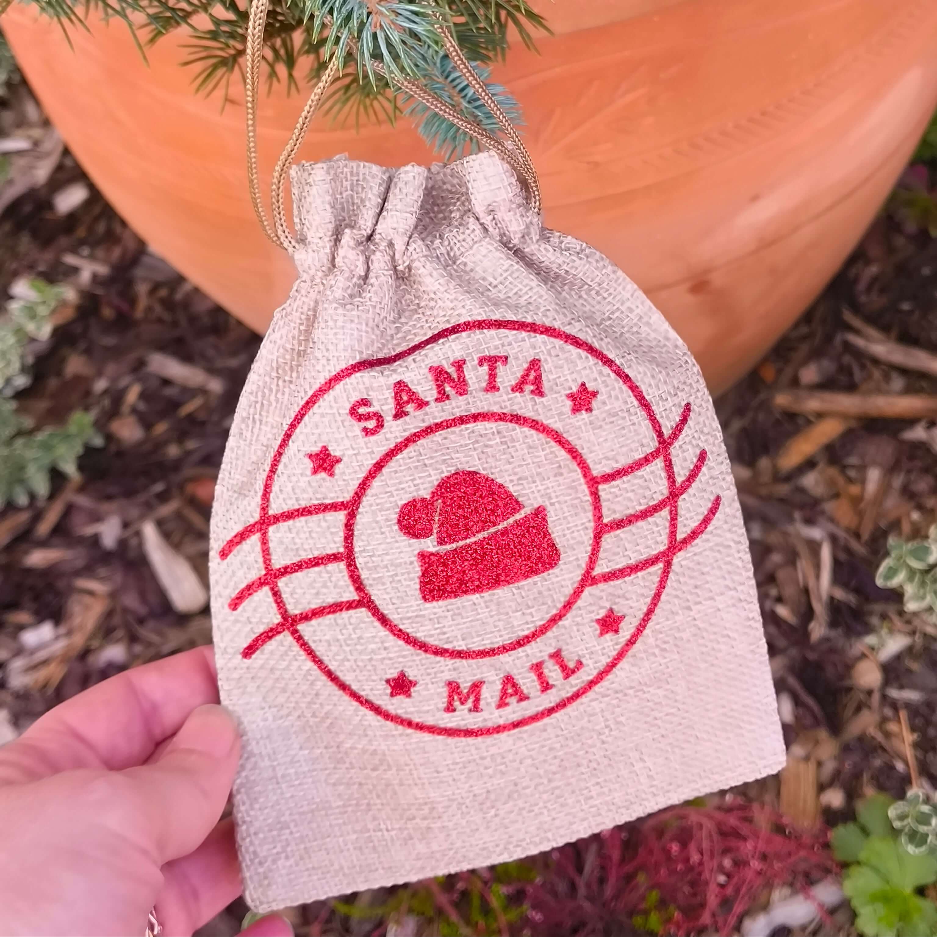 Hand holding a small hessian Santa Mail gift bag with red design, outdoors near a Christmas tree