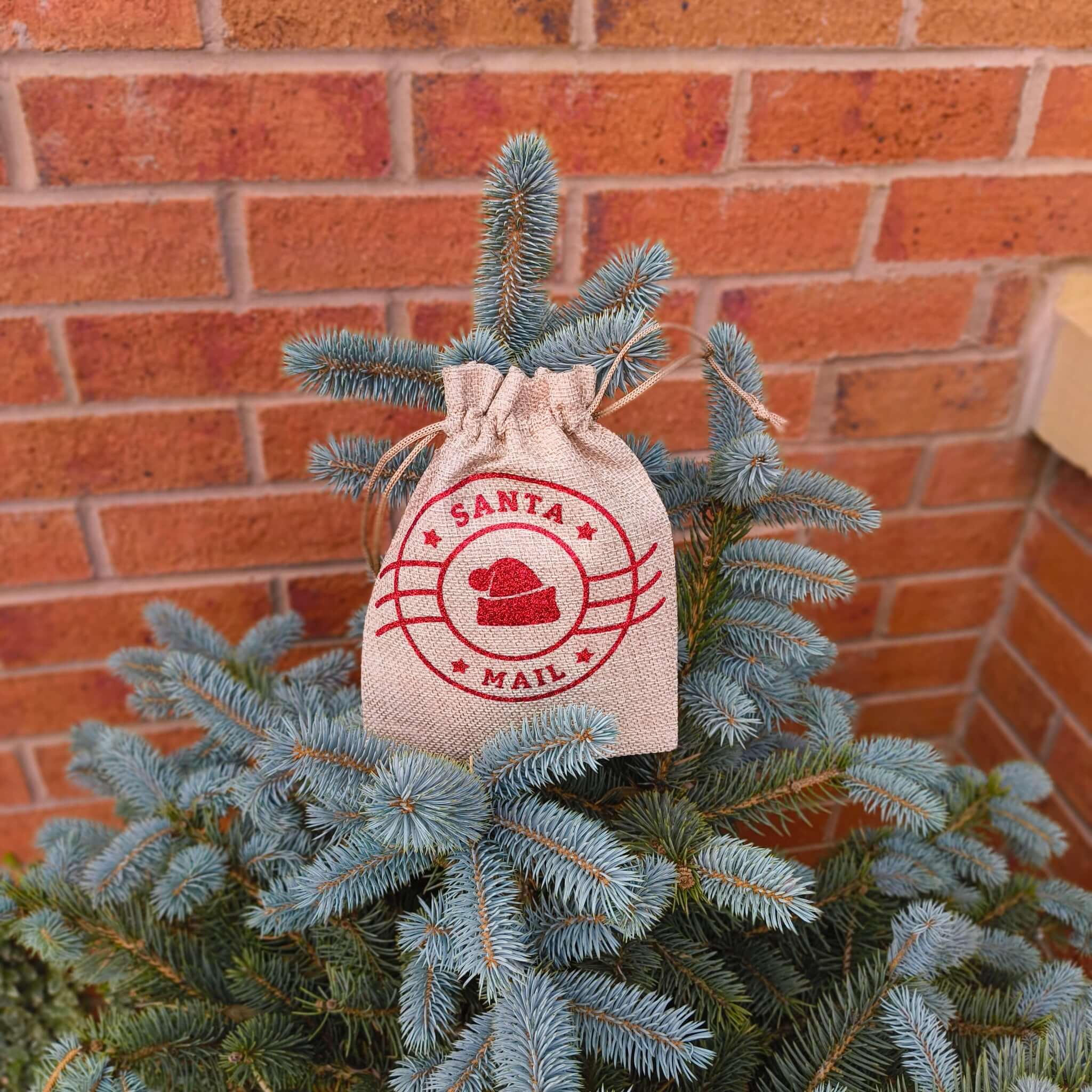 Santa Mail hessian bag with red festive print, hanging on Christmas tree branches near brick wall.