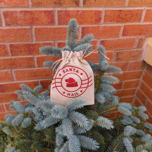 Santa Mail hessian bag with red festive print, hanging on Christmas tree branches near brick wall.