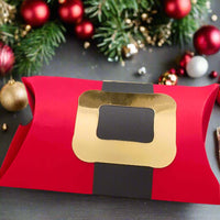 Handmade red Christmas pillow gift box styled like Santa’s suit with a gold foil buckle and black belt, displayed on a dark wooden surface with festive decorations including red and gold baubles, fir branches, and scattered ornaments in the background.