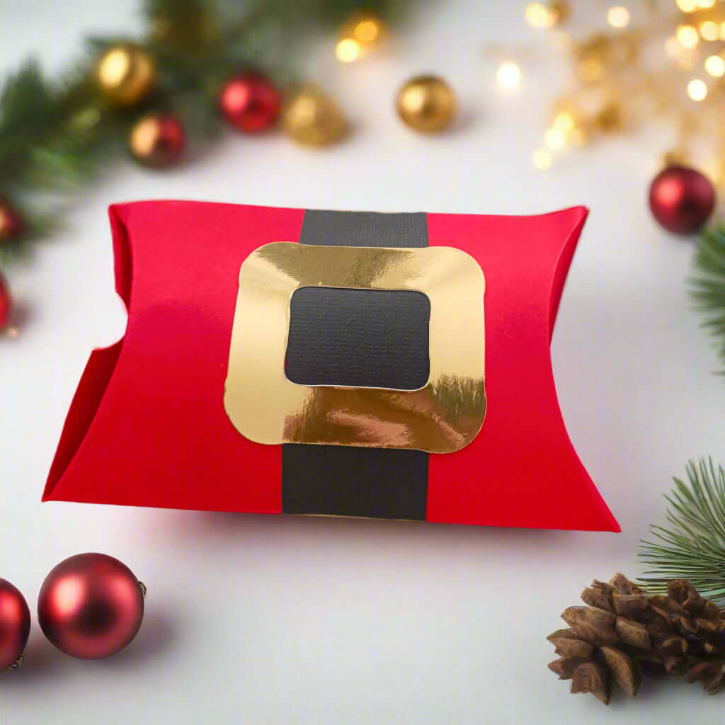 Red Santa-themed pillow gift box with a gold buckle and black belt design, surrounded by festive Christmas decorations including pinecones, red and gold baubles, fir branches, and warm fairy lights on a soft white background.