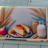 Handmade Shavuot greeting card featuring two slices of cheesecake, a milk jug, a pomegranate, wheat stalks, nuts, and a prayer book, displayed on a light background.
