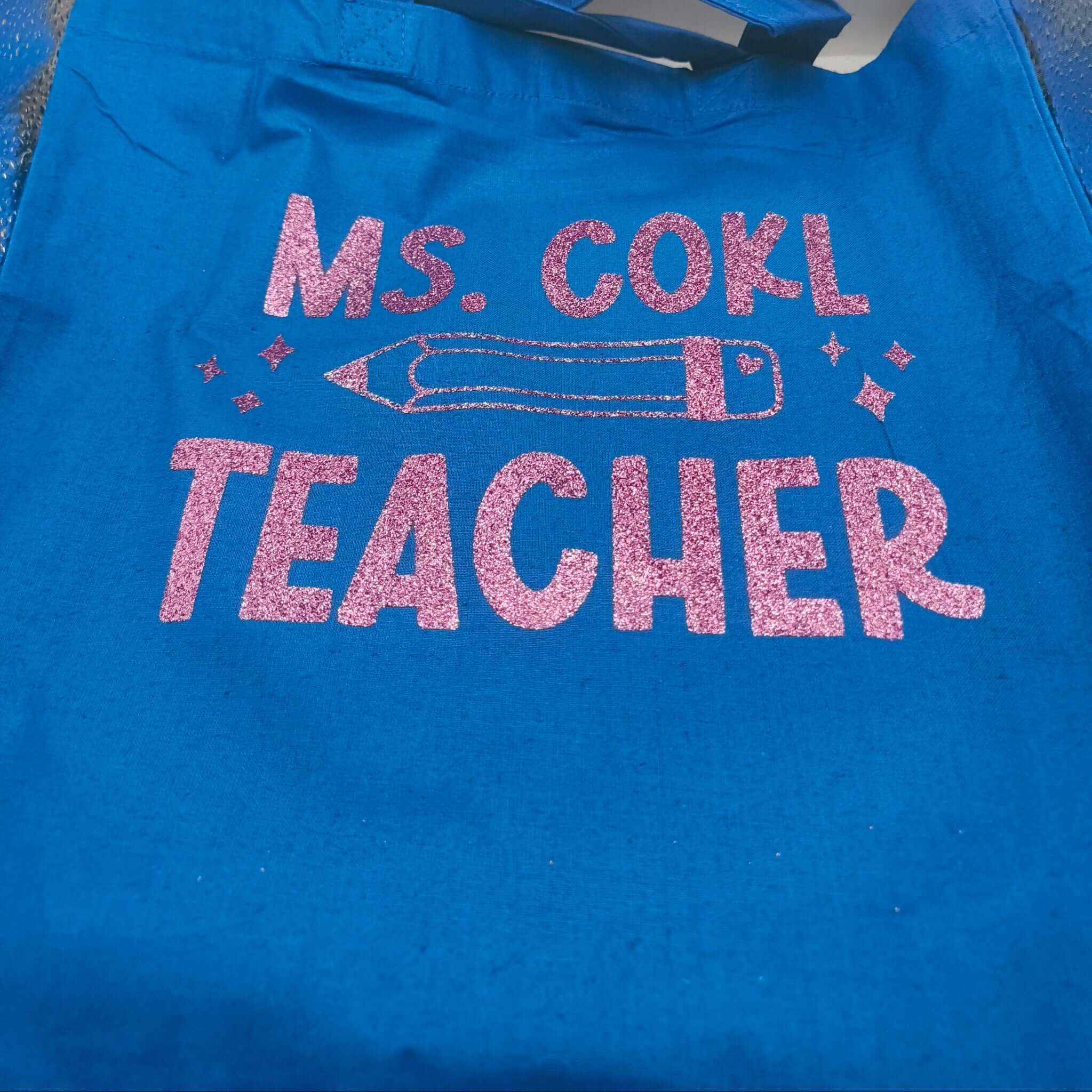 Flat lay of a blue tote bag with pink glitter text "Ms. Cohl Teacher" and pencil design, displayed against a neutral background.