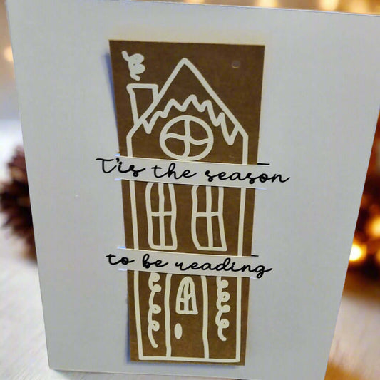 Handmade Christmas card featuring a brown gingerbread-style house design with white icing details. Across the card, black cursive text reads: '’Tis the season to be reading'. The background is softly lit with warm, festive lights.