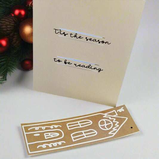 Handmade Christmas card displayed upright with black script text reading '’Tis the season to be reading'. A matching brown gingerbread-style house bookmark with white detailing lies in front of it. A festive pine branch with red and gold baubles is in the background.