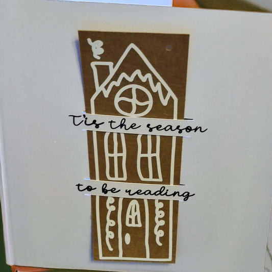Handmade Christmas card featuring a tall gingerbread-style house in white on a brown background. Across the house, two banners read: '’Tis the season to be reading' in black cursive font.